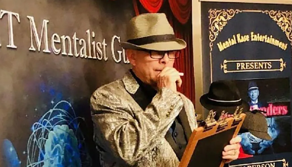 A performer in a glittery jacket and fedora holds a clipboard and appears deep in thought at an event presented by Mentalist G
