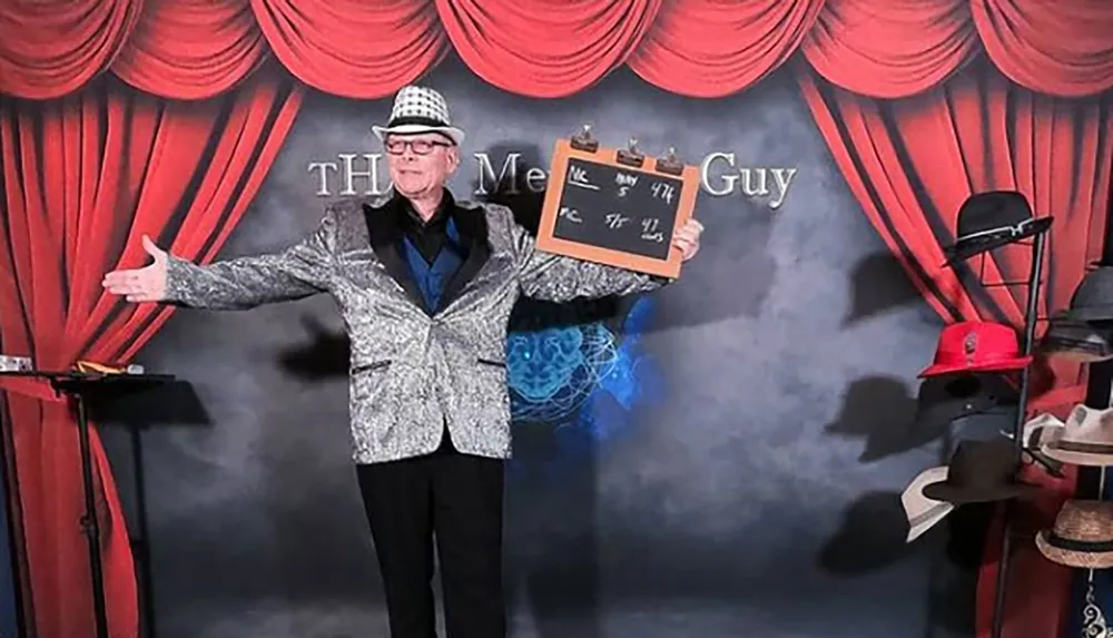 A person in a shiny jacket and checkered hat is holding a clapperboard and gesturing with an open arm in front of a backdrop with red curtains and the text The Mental Guy