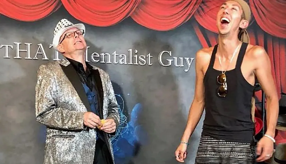 Two people are standing onstage with contrasting expressions and attires one in a shiny silver jacket looking amused and the other in a casual sleeveless top laughing exuberantly against a backdrop with the words That Mentalist Guy