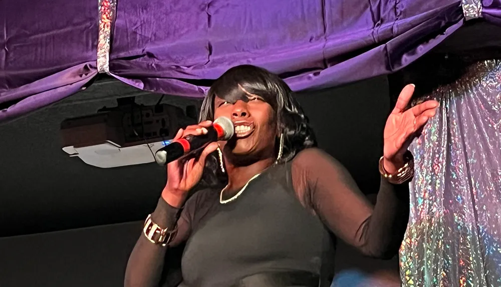 A person is singing passionately into a microphone gesturing expressively with one hand against a background of purple drapery and a shimmering fabric