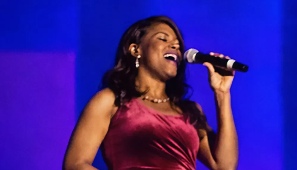 A woman in a burgundy dress is passionately singing into a microphone on stage with a blue background