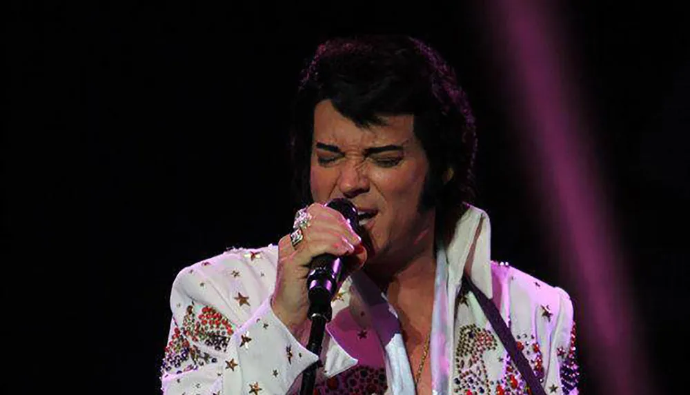 A performer dressed in a white embellished jumpsuit is singing into a microphone on stage with a focused expression