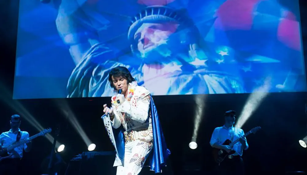 A performer dressed as Elvis Presley sings on stage with a band while a large image of the Statue of Liberty is projected in the background