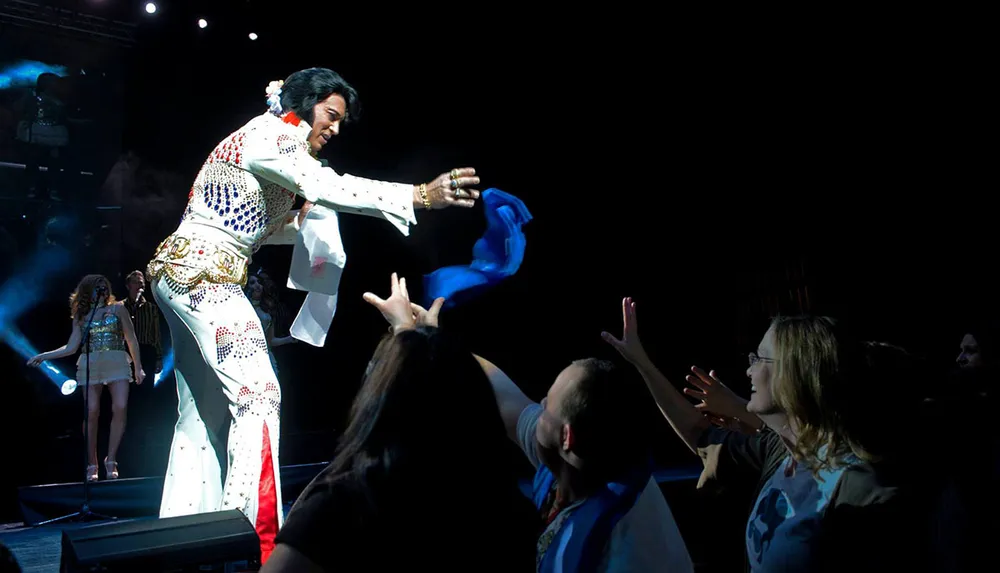 An Elvis impersonator in a jumpsuit is throwing a scarf to an excited audience