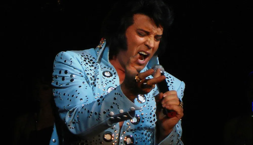 A person dressed in a sparkling blue jumpsuit is singing passionately into a microphone on stage