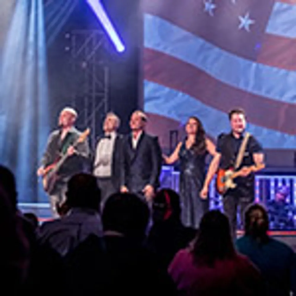 A band performs on stage in front of an audience with an American flag projected in the background