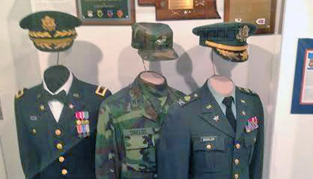 The image displays a collection of military uniforms and hats presumably from the United States showcased on mannequins with a backdrop of medals and commendations in what appears to be an exhibit or a personal collection