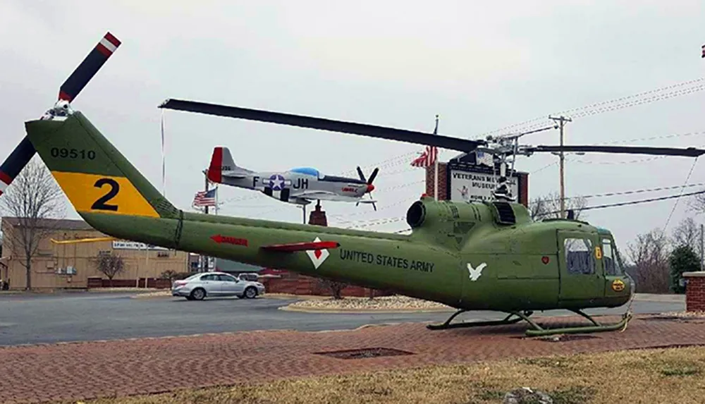 An olive green US Army helicopter is displayed in front of a museum with a vintage plane in the background and an American flag flying above