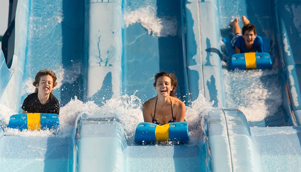 Three people are joyfully sliding down a water slide on blue mats
