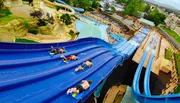 Three people are enjoying a sunny day at a water park, sliding down in a large inflatable ring with big smiles on their faces.
