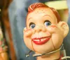 The image shows a close-up of a vintage-looking toy with a smiling freckled face and blue eyes accompanied by blurred background figures