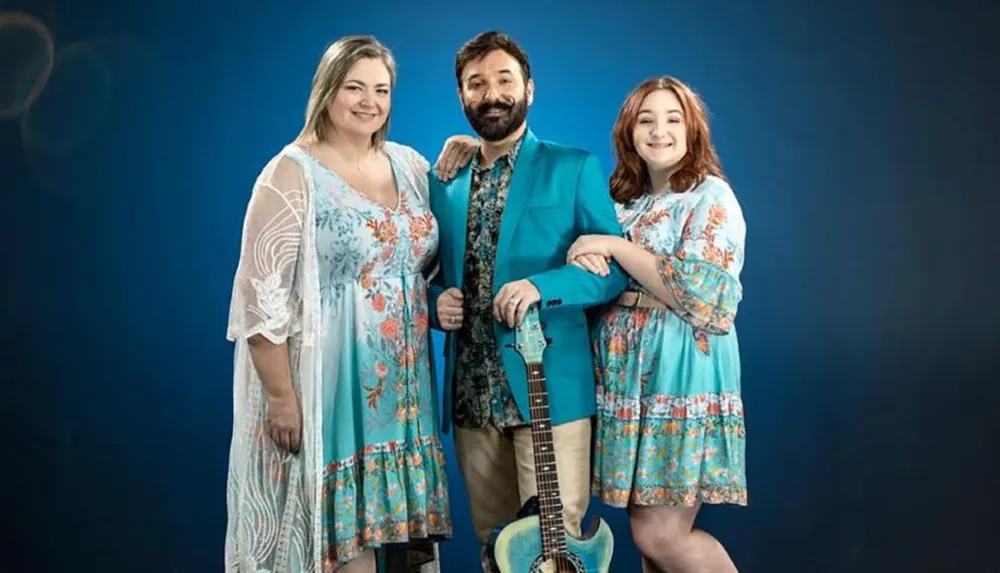 Two women and a man with a guitar are posing cheerfully against a blue background possibly suggesting a musical or familial theme