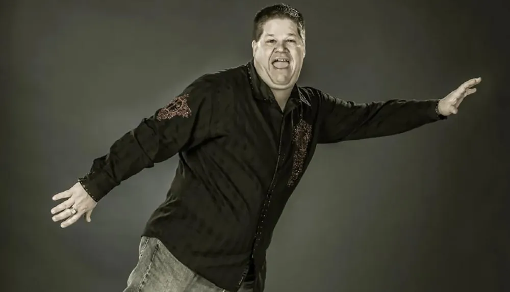 A man with a jubilant expression is extending his arms as if balancing or gesturing enthusiastically wearing a decorated black shirt and jeans set against a grey background