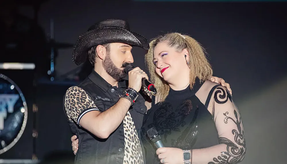 A man wearing a cowboy hat is singing into a microphone closely to a smiling woman who is also holding a microphone possibly performing a duet on stage