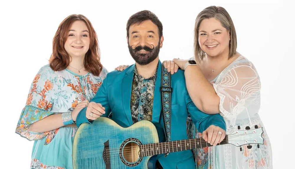 Three smiling people are posing together with the central figure holding a blue guitar and wearing a bright blue suit flanked by two women in patterned dresses on a white background