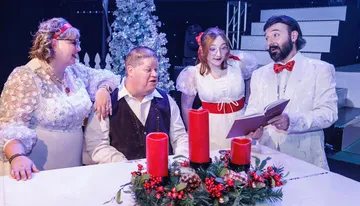 A group of people dressed in festive attire are gathered around a table with a Christmas wreath and candles, singing or reading together in a holiday setting.