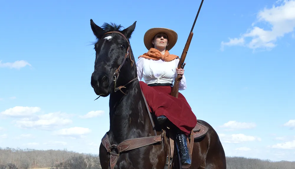 A person dressed in historical attire is mounted on a black horse holding a long rifle under a blue sky with clouds