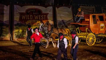 Performers in Wild West attire are present in front of a stagecoach, with one man smiling and twirling a lasso, evoking the atmosphere of a classic western show.