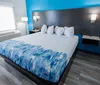 The image shows a modern hotel room with a large bed featuring a blue and white patterned comforter set against a wood-paneled wall and accented by vibrant blue walls