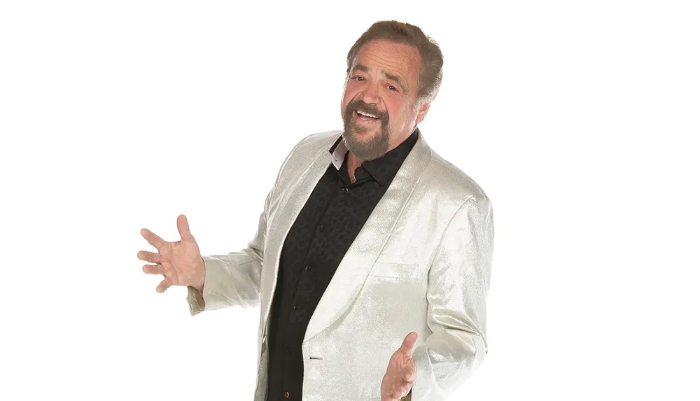 A man with a beard is smiling and gesturing with his hands while wearing a shiny silver jacket over a black shirt