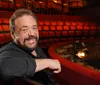 A smiling man is seated in an empty theater with red seats and a stage in the background