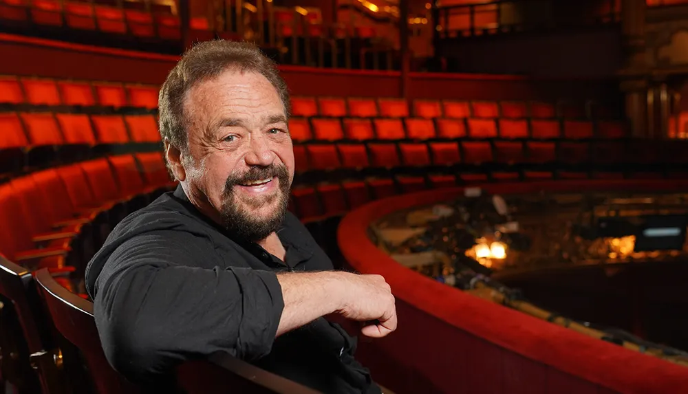 A smiling man is seated in an empty theater with red seats and a stage in the background