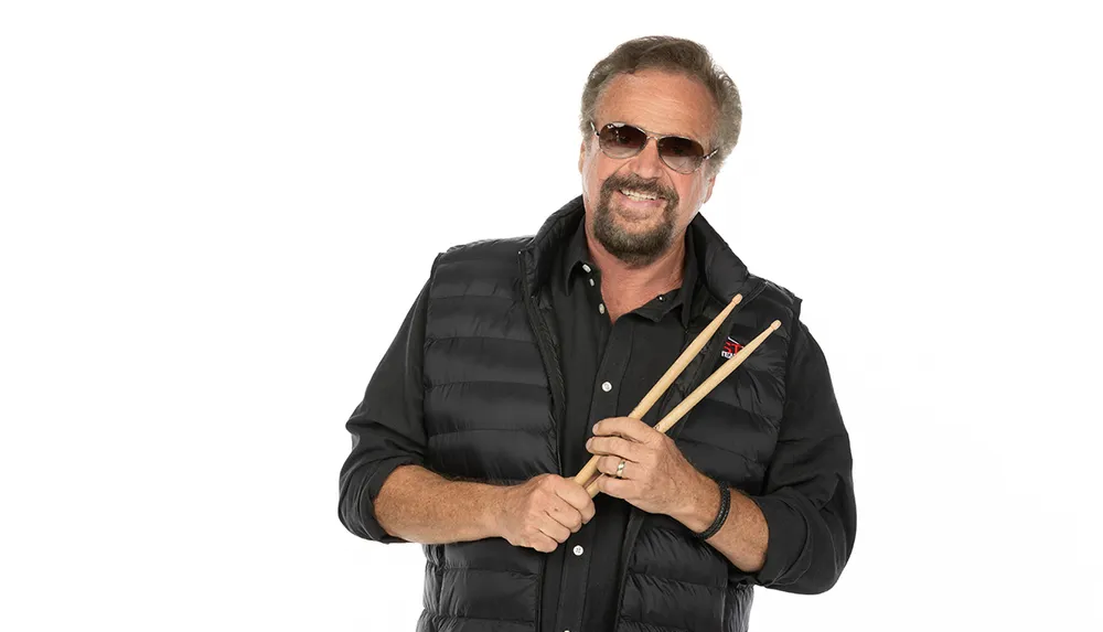 A smiling man with sunglasses and a beard is wearing a black vest holding a pair of drumsticks against a white background