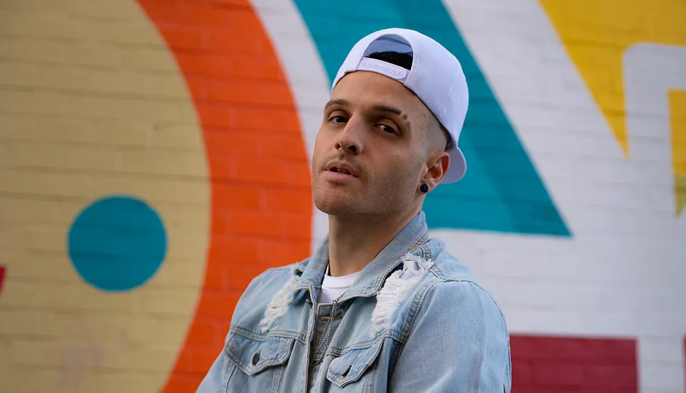 A person wearing a white cap and a denim jacket stands in front of a colorful mural with geometric shapes