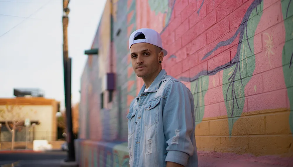 A person is standing in front of a colorful graffiti wall wearing a white cap and denim jacket with a casual yet thoughtful expression