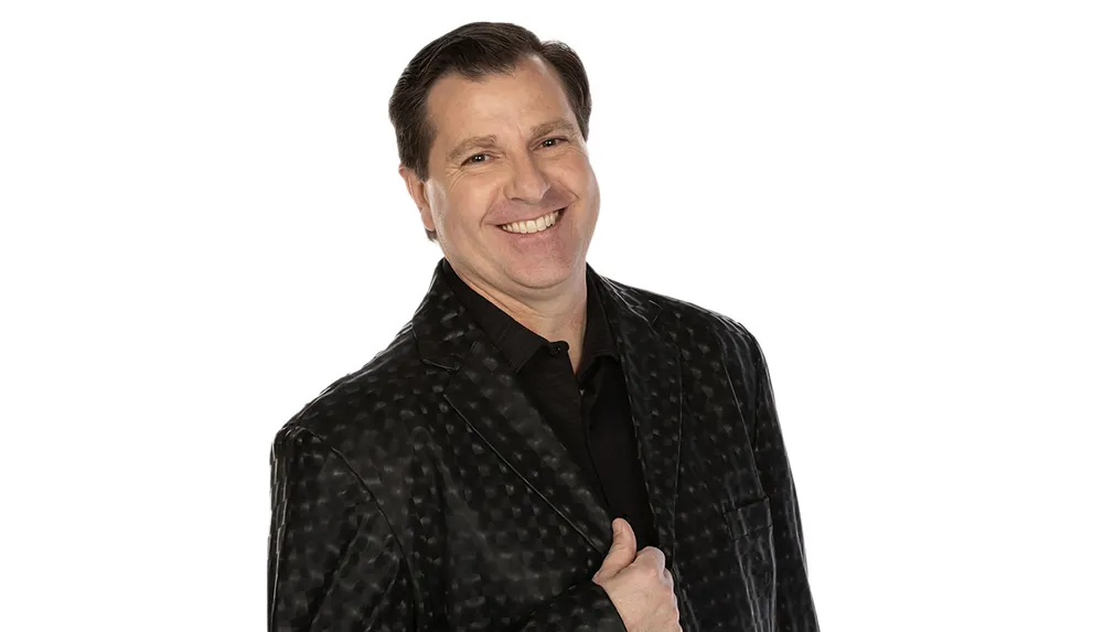 A smiling man in a dark patterned jacket with his arms crossed stands against a white background