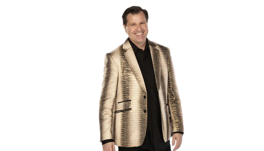 A man is smiling at the camera wearing a black shirt and a shiny gold blazer against a white background