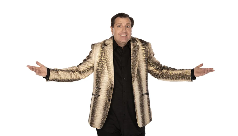 A smiling person is wearing a shiny gold blazer with arms outstretched seemingly expressing a welcoming or questioning gesture against a white background