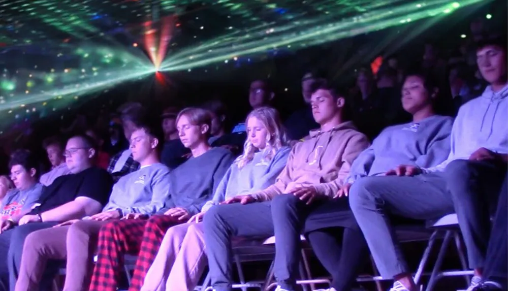 A group of people appears to be in a trance-like state against a backdrop of colorful laser lights possibly during a hypnosis show or a similar event