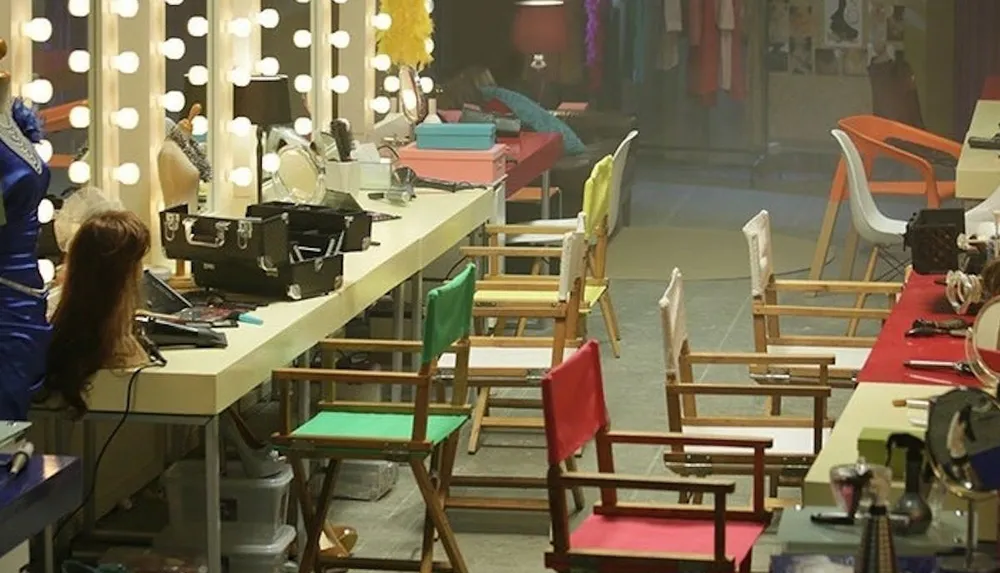 The image shows a brightly lit dressing room with a large mirror surrounded by light bulbs a makeup table lined with various cosmetics and tools colorful chairs placed in front and costumes displayed in the background