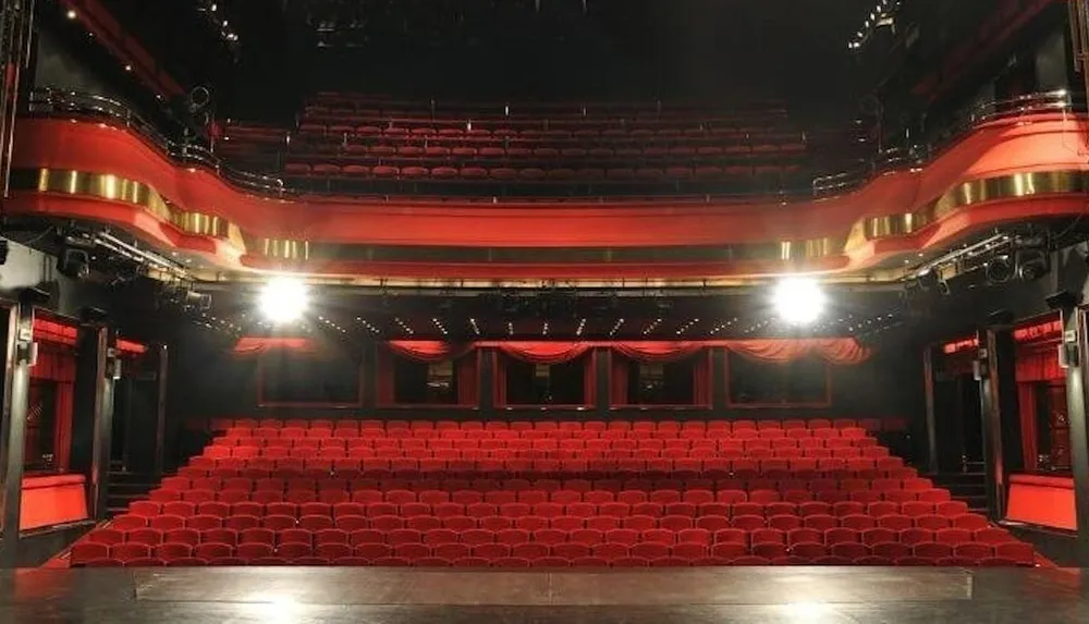 The image shows an empty theater auditorium with red seats and multiple levels of balconies facing a stage conveying a sense of anticipation for a performance