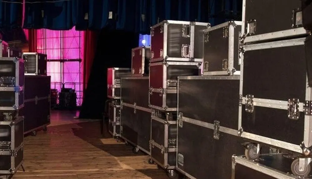 A row of road cases typically used for transporting stage equipment is positioned on a wooden floor backstage with colorful stage lighting visible in the background