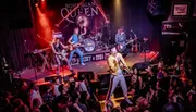 A band is performing on stage in front of an enthusiastic audience, with the lead singer positioned prominently at the forefront, evoking the style of a vintage rock concert.