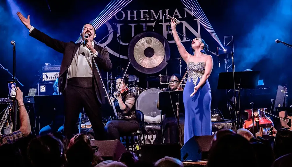 Two vocalists passionately perform on stage with a backing band and an audience in the foreground with the bands name BOHEMIAN QUEEN displayed on the backdrop