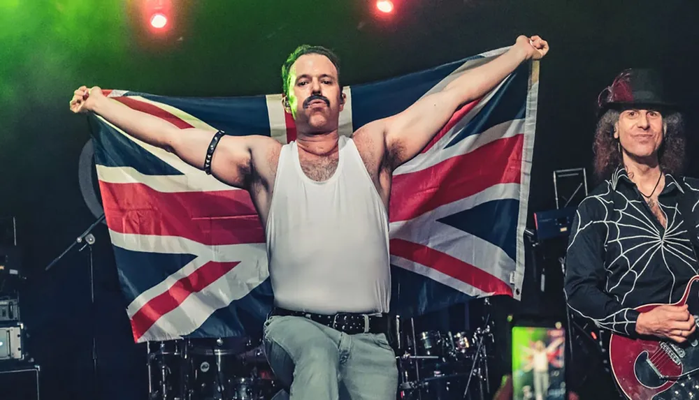 A performer onstage emulates a well-known pose often associated with a famous rock singer holding up a Union Jack flag with a guitarist beside him