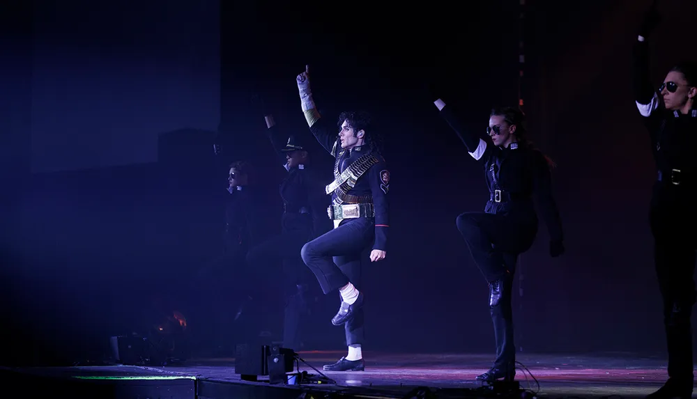 A performer emulating the iconic style of Michael Jackson is dancing onstage with backup dancers in matching dark outfits