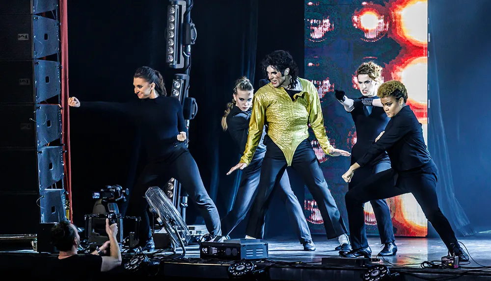 A performer in a gold shirt and black pants is dancing on stage with a group of dancers in black attire against a colorful backdrop while a camera operator captures the scene from the wings