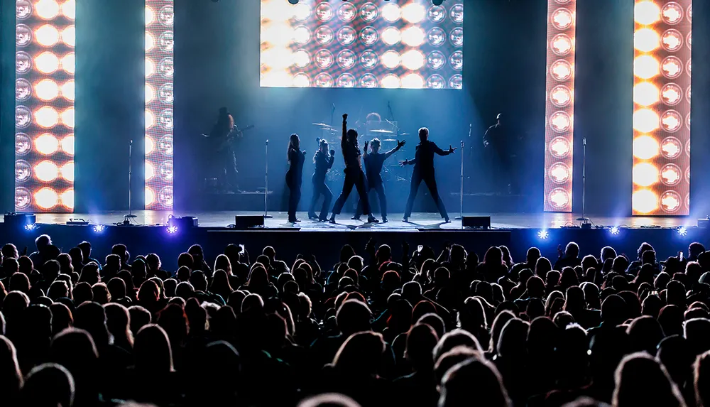 A silhouette of musicians and dancers performing on stage backlit by bright lights with an audience in the foreground