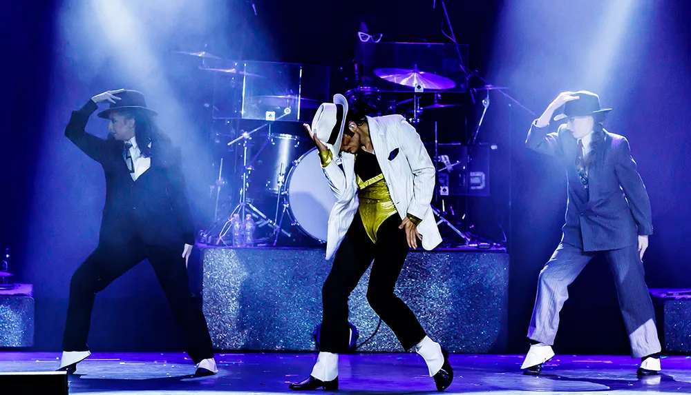 Three performers onstage exhibit a dynamic dance pose reminiscent of Michael Jacksons iconic style with the central figure donning a white jacket and gold belt
