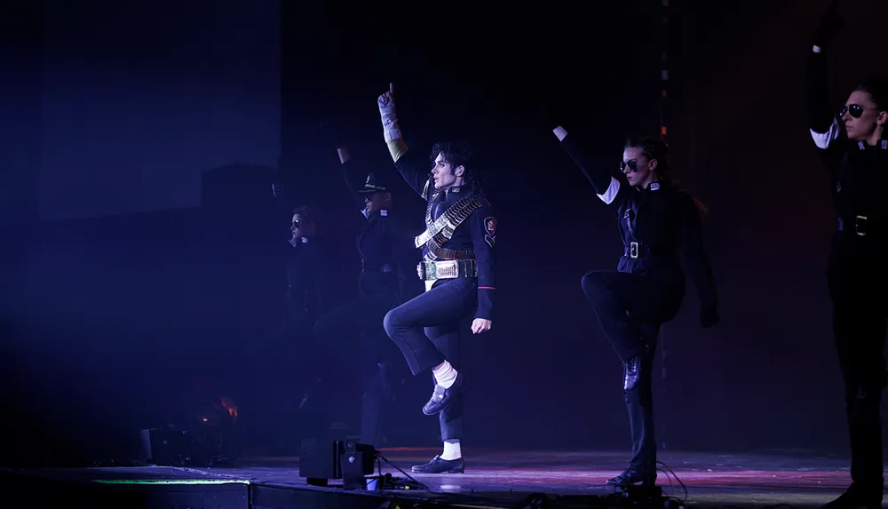 A group of performers dressed in military-style attire are energetically dancing on stage with one leading in the center