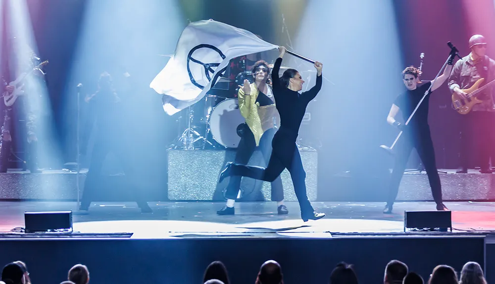 A dynamic stage performance features a band with a person energetically waving a large flag