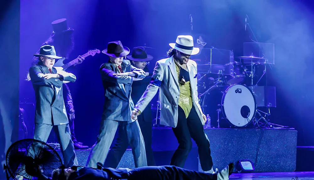 A group of performers in suits and hats are dancing energetically on stage with a drummer in the background