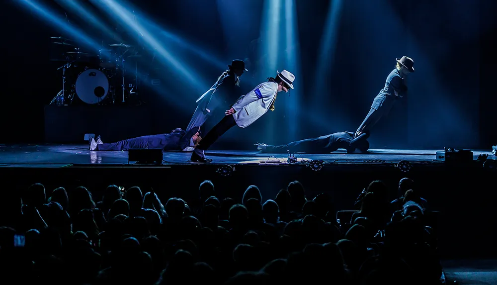 Dancers dressed in dark suits and white fedoras perform a gravity-defying lean on a dimly lit stage with an audience watching