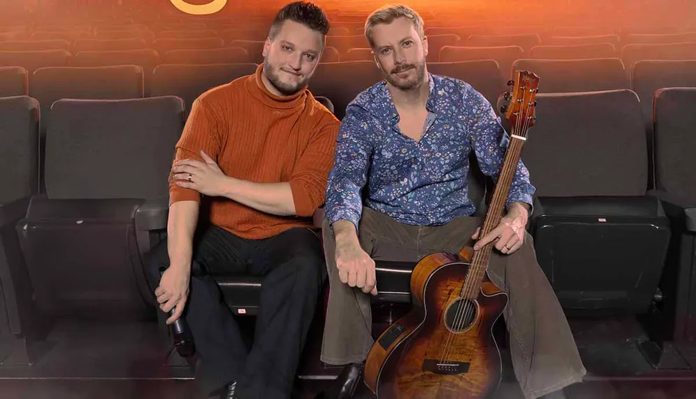 Two men are sitting together in a theater one holding an acoustic guitar with a relaxed and friendly demeanor