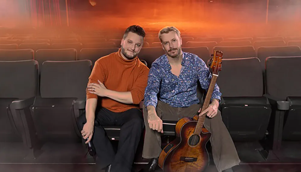 Two men are sitting in theater seats one holding a guitar with a warm smoky background