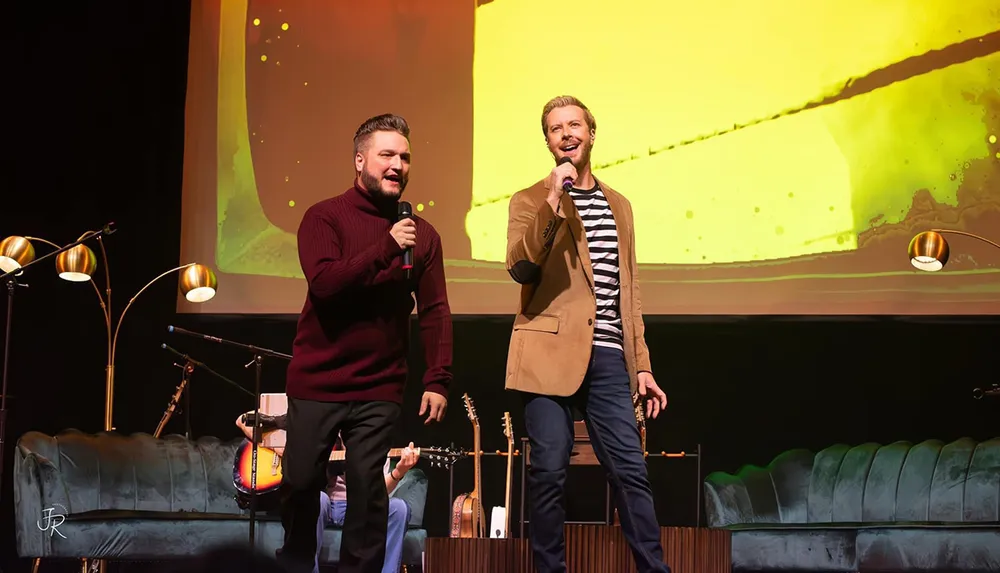 Two men are enthusiastically performing on stage with microphones surrounded by musical instruments and a vibrant background